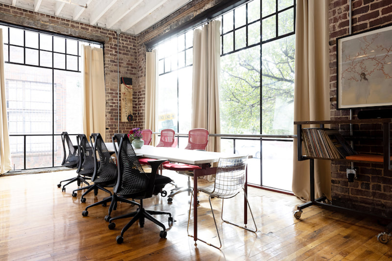 View of conference table on hardwood flooring with 7 rolling office chairs, a tall vintage industrial standing desk, large loft windows with diffusing and black out curtains 