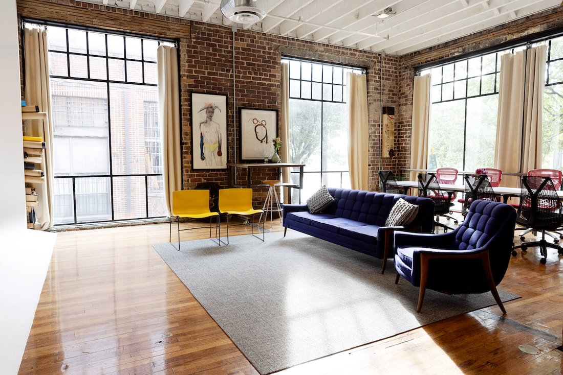 Beautiful and colorful perspective of the sitting area. There is hardwood flooring throughout with a couch and many different types of chairs, a standing rustic vintage desk, unique paintings on the walls and a conference table with 7 rolling office chairs. The Cyclorama wall is peeking out from the left and the entire room has daylight coming through the large 13 foot loft windows 