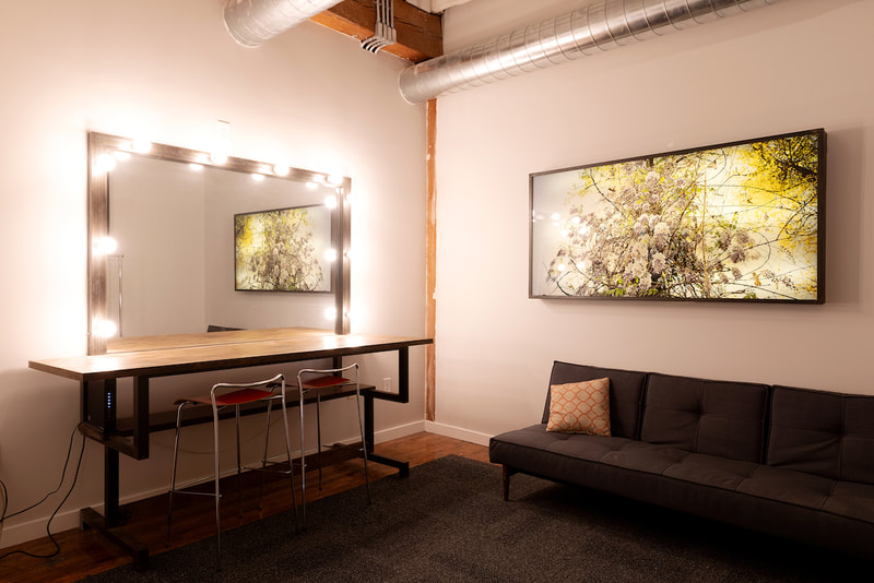 A room for makeup and dressing with a large mirror framed with big lightbulbs, sitting on a tall  bar hight industrial wooden desk and bar stools for makeup application. On the adjacent wall there is a dark grey couch sitting below a large, modern, glowing light box painting.